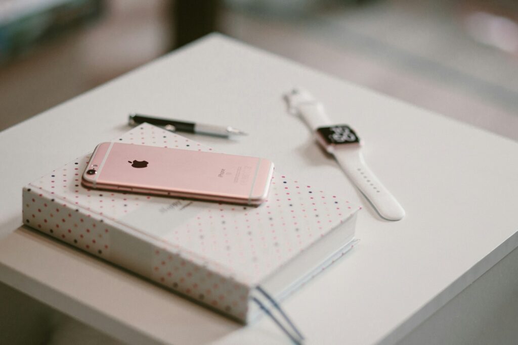 iphone rosa encima de un libro con un bolígrafo y un reloj encima de una mesa blanca